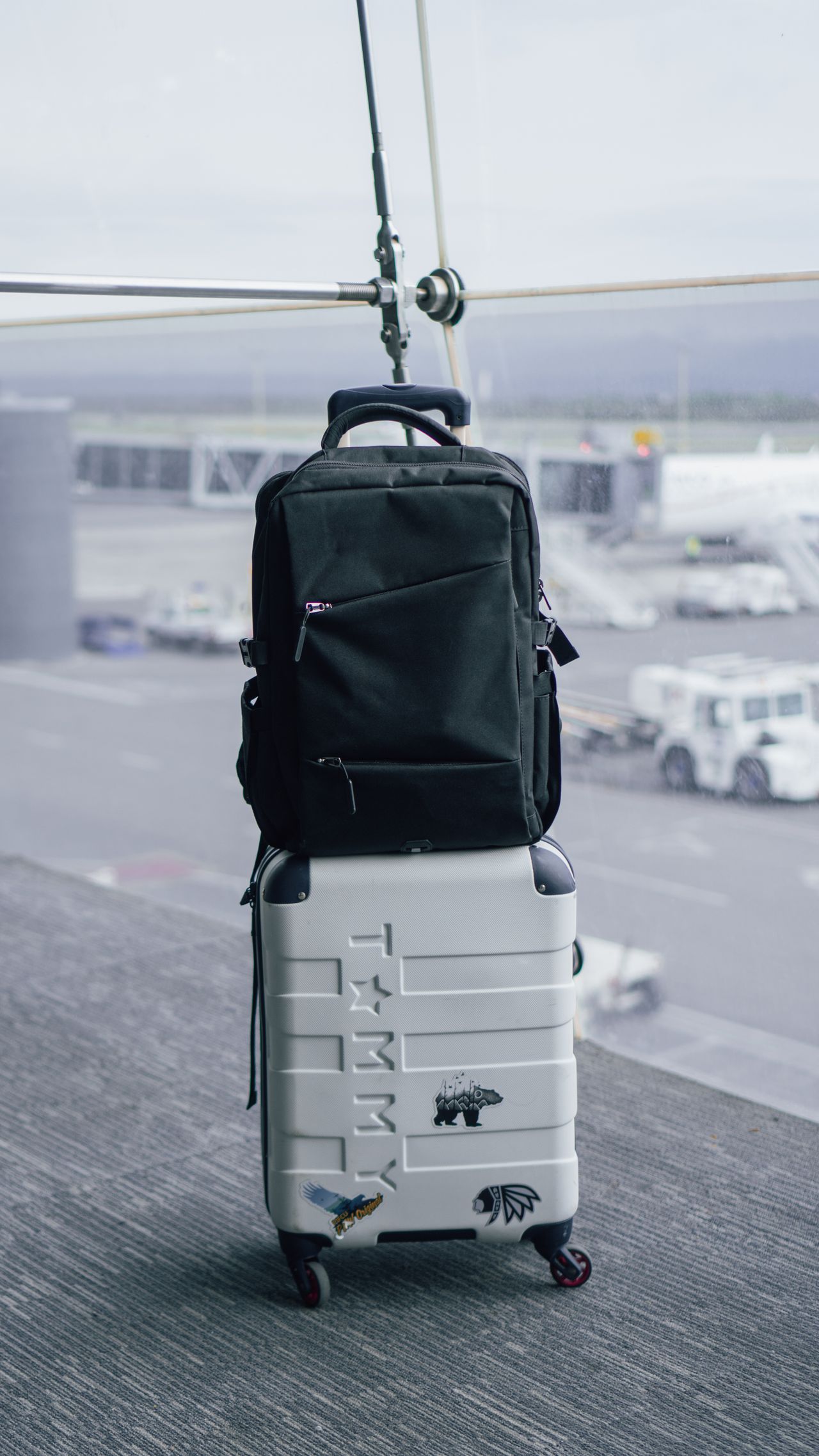 Black backpack resting on a white wheeled suitcase beside airport terminal windows